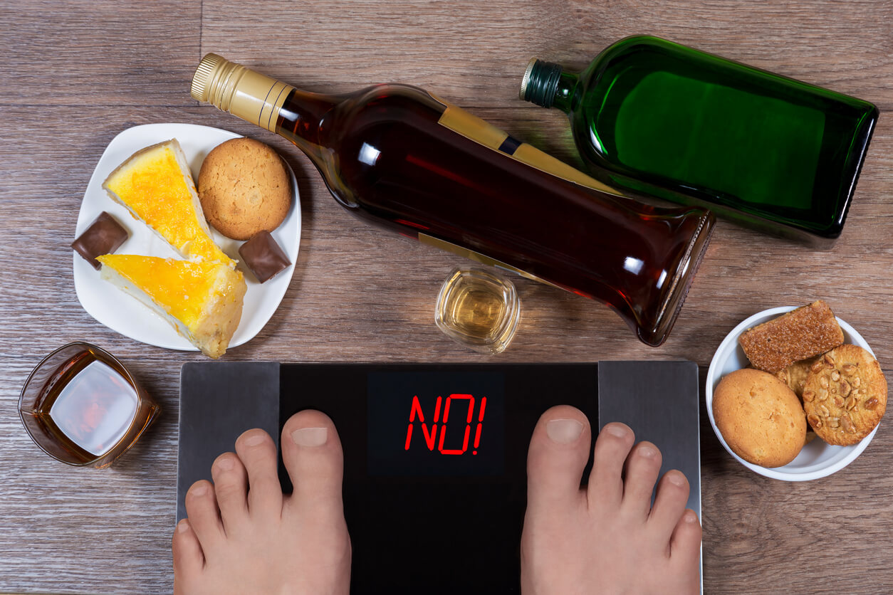 Male feet on digital scales with word no on screen. Bottles and glasses of alcohol, plates with sweet food. Concept of consequences of unhealthy lifestile. Morning after party.